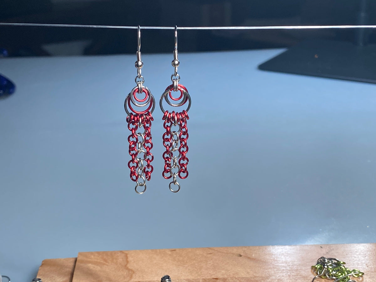 A pair of handmade stainless-steel dangle earrings with red and silver chainmail-style fringe, hanging from a thin wire against a light blue-gray background.