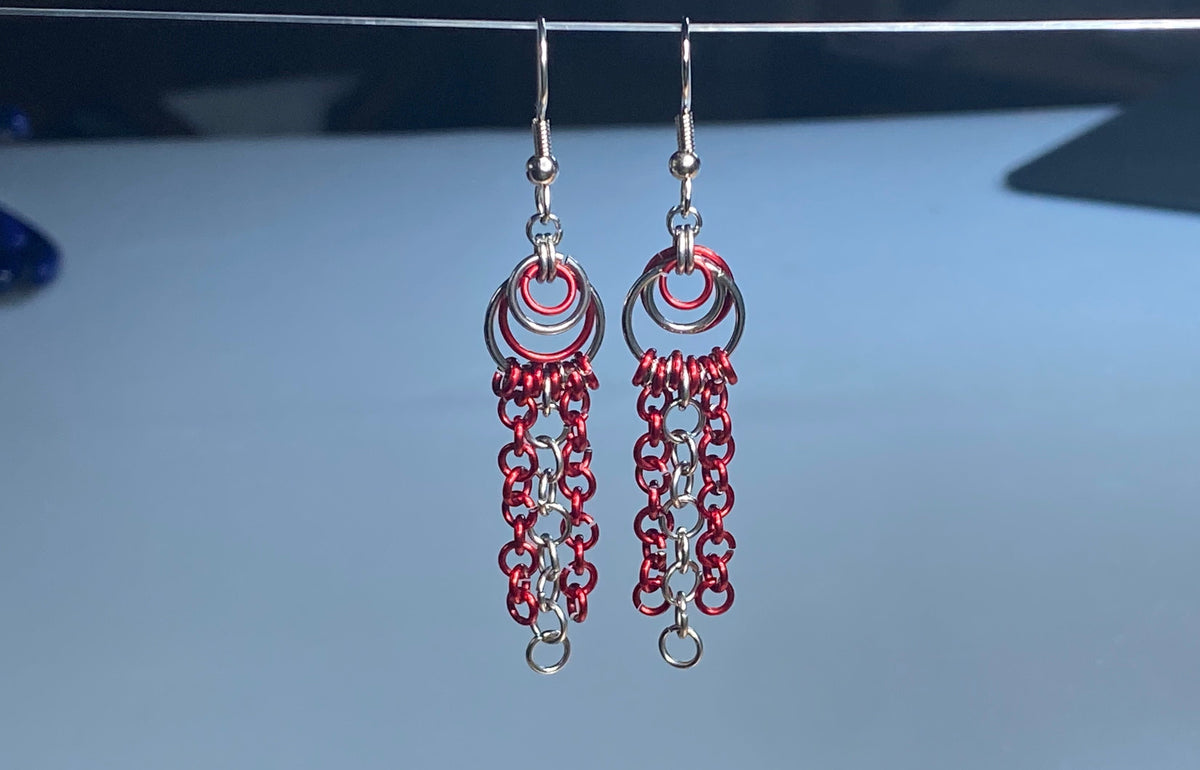 A pair of handmade stainless-steel dangle earrings with red and silver chainmail-style fringe, hanging from a thin wire against a light blue-gray background.