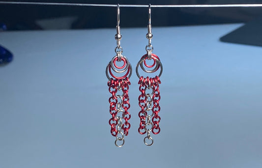 A pair of handmade stainless-steel dangle earrings with red and silver chainmail-style fringe, hanging from a thin wire against a light blue-gray background.