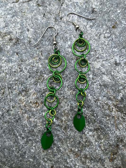 A pair of handmade dangle earrings with multiple shades of green graduated jump rings, large to small complete with dark green aluminum pendants displayed on a natural gray stone with silver streaks and flecks.