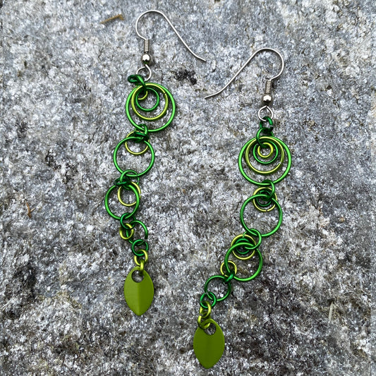 A pair of handmade dangle earrings with multiple shades of green graduated jump rings, large to small complete with light green aluminum pendants displayed on a natural gray stone with silver streaks and flecks.